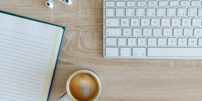 A keyboard, notebook, headphones and cup of coffee sitting on a desk