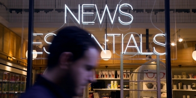 The top of a man's head walking past a shop with a neon sign that reads news essentials