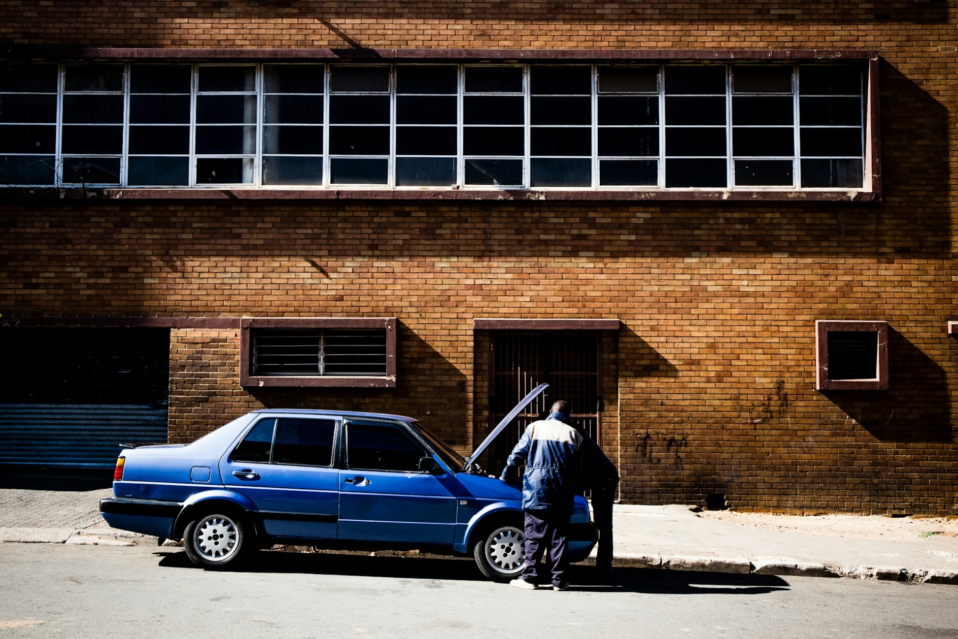 Two men looking under the hood of a blue car, parked on the street in front of a brick building