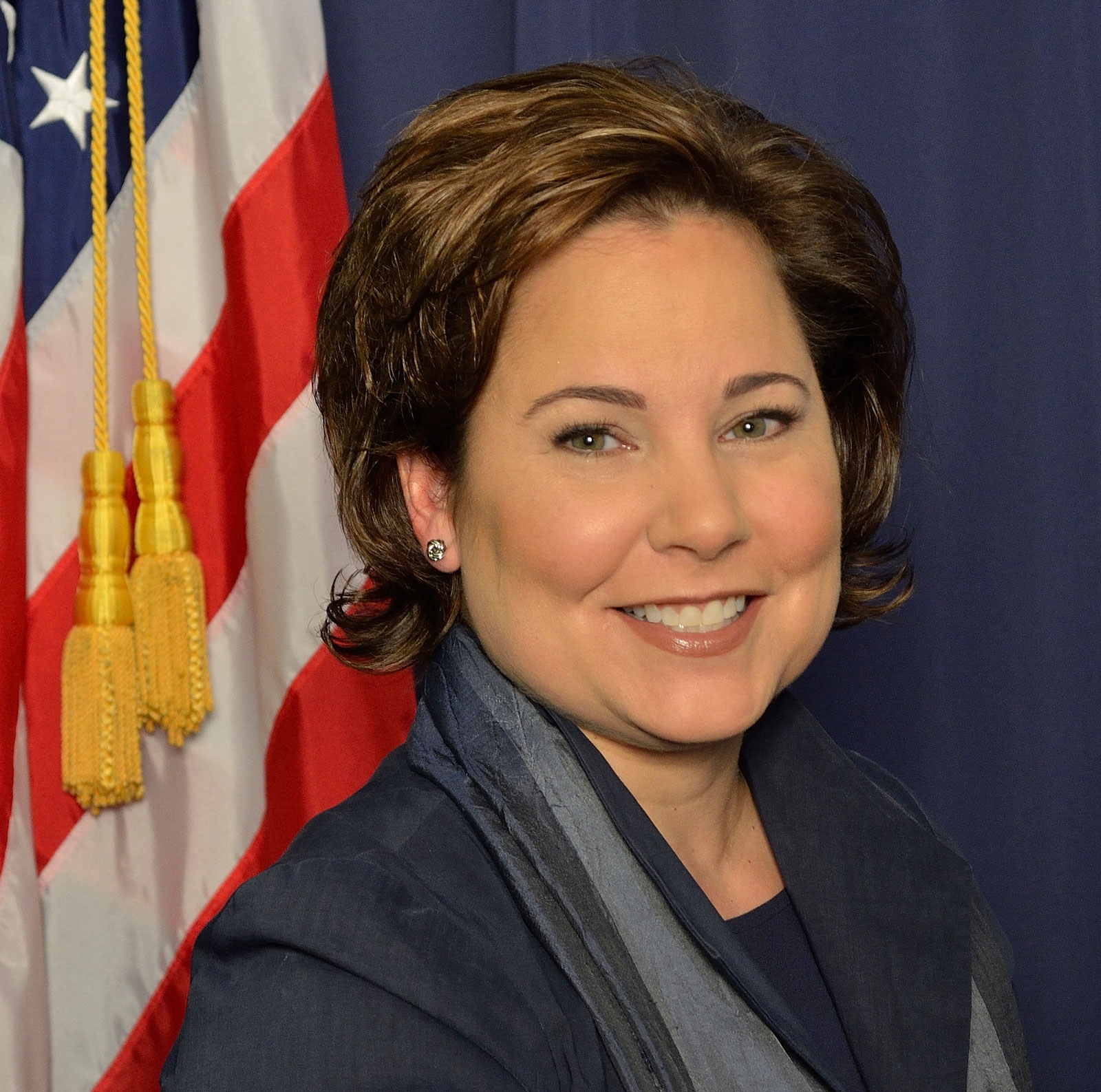 Headshot of a middle-age white woman, with short brown hair, wearing a business suit and smiling while standing in front of an American flag