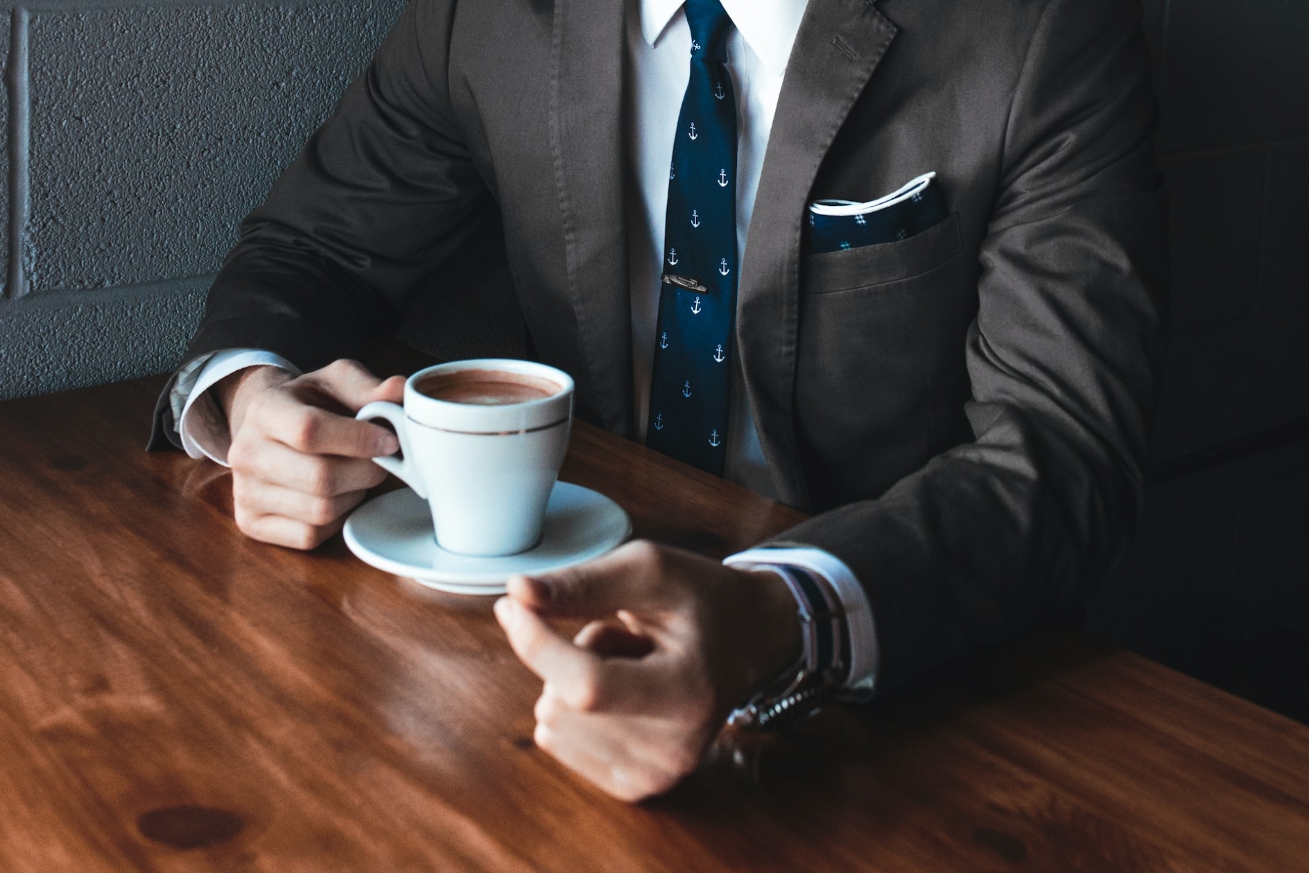 A man wearing a suit, sitting at a wooden table, drinking coffee