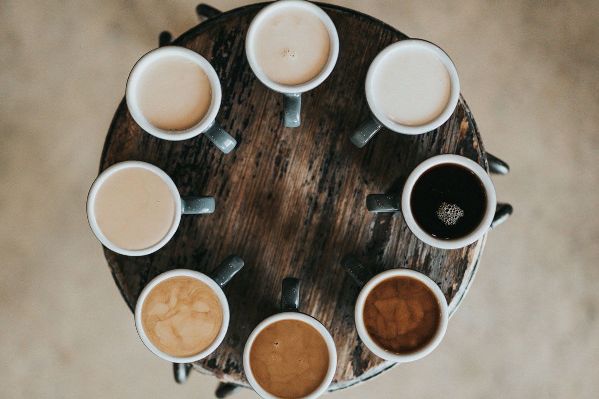 eight cups of coffee sitting on a stool, each with a different amount of creamer, creating a spectrum from black to white
