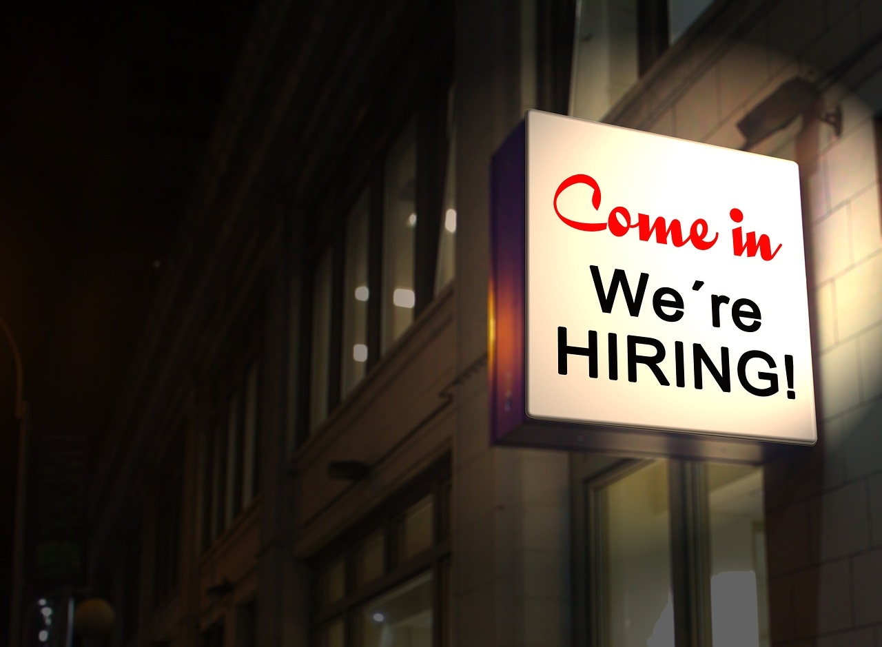 Photo of an illuminated sign on the side of a building that reads Come in We're Hiring!