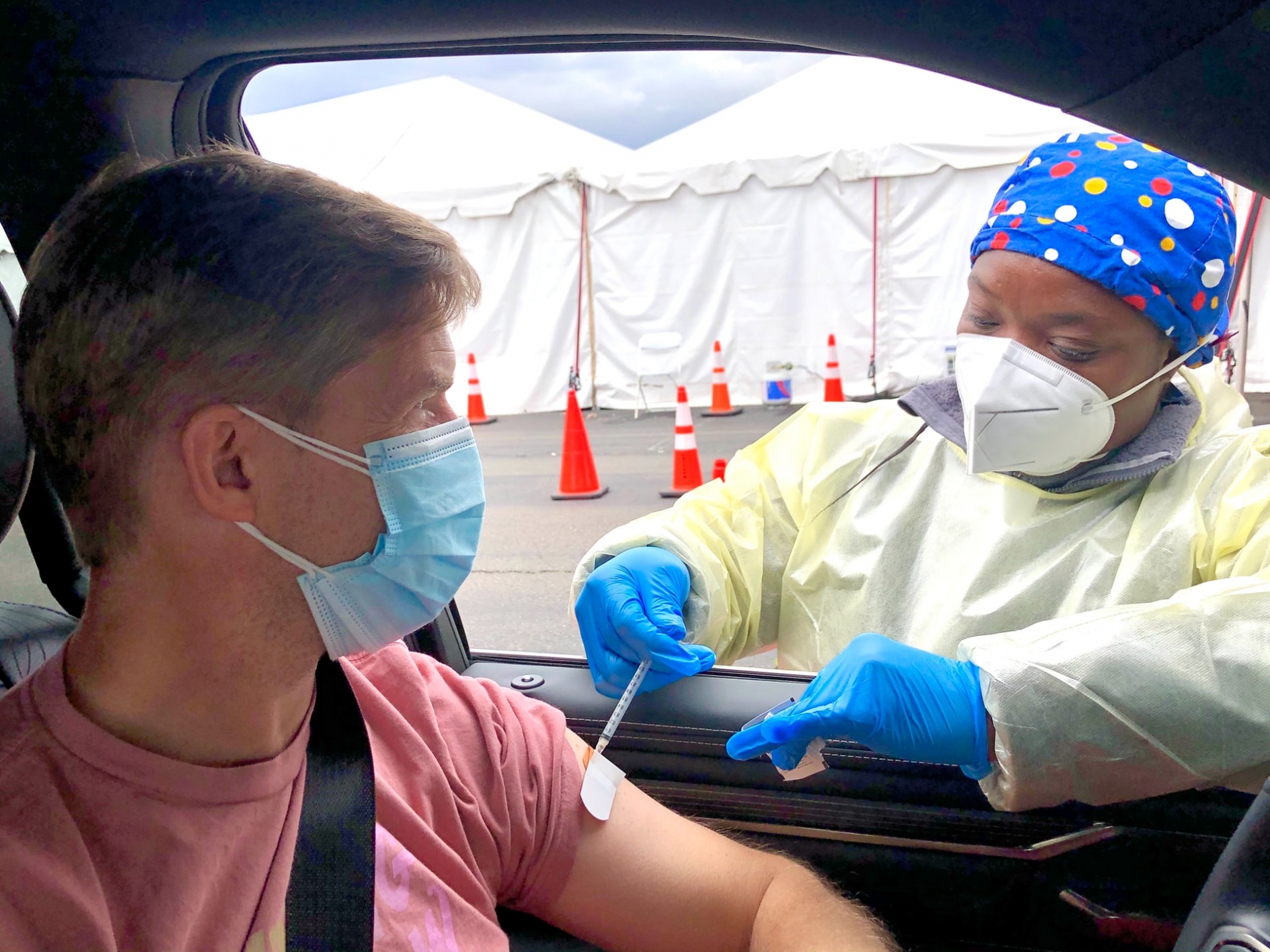 A man wearing a mask, sitting in his car, getting a vaccine shot in his left arm through the open window from a nurse wearing PPE