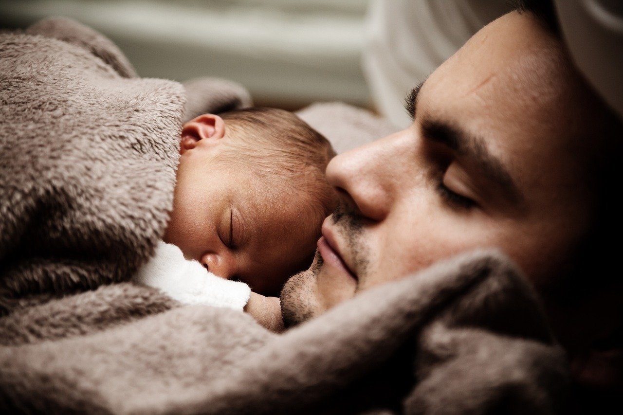 Closeup of a man's face, reclining, eyes closed, with a tiny baby sleeping on his chest, wrapped in a blanket