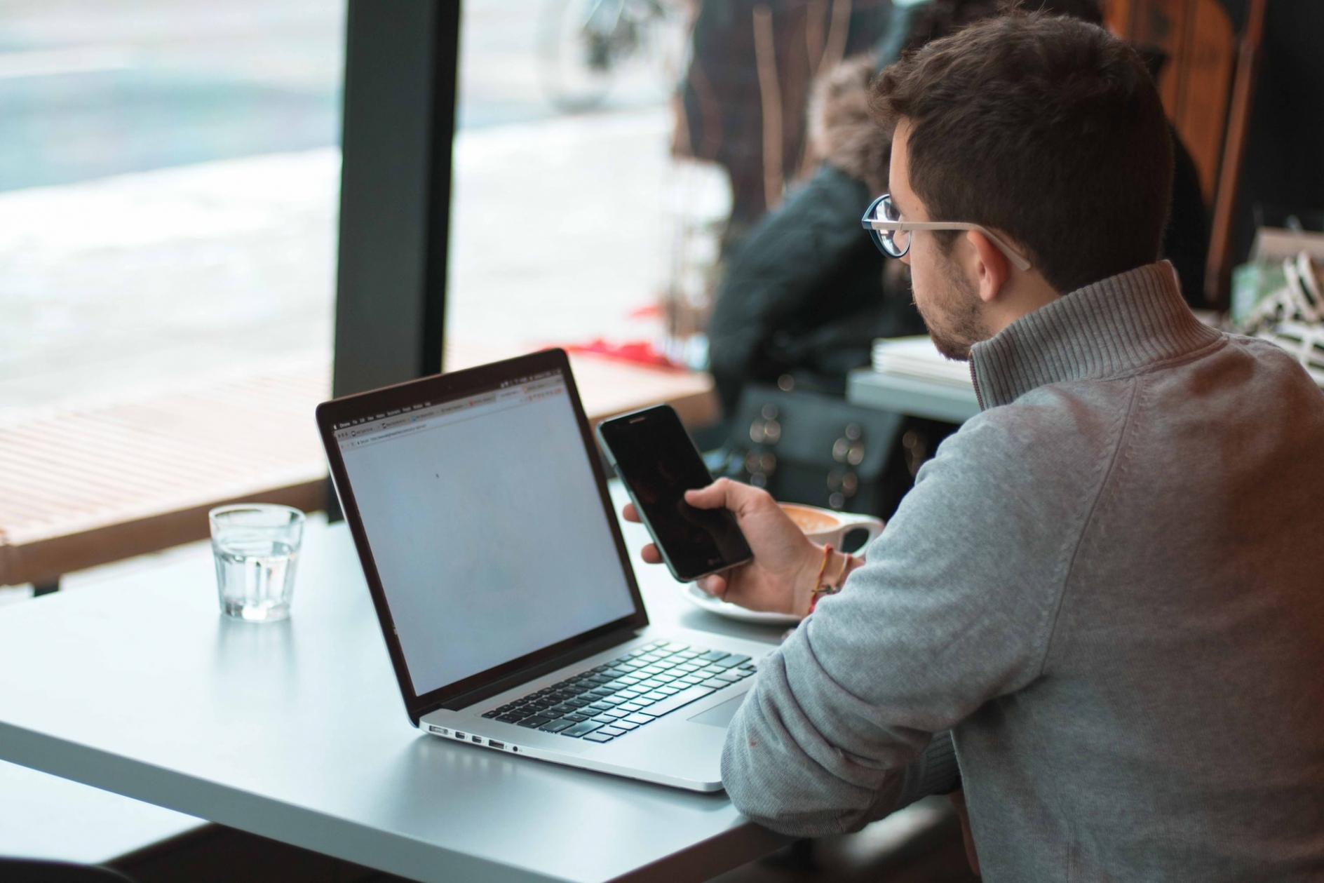 A man wearing glasses and a grey sweater sits at a table in a cafe, looking at his smartphone, with a notebook computer open in front of him