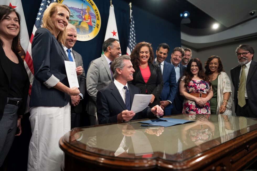 California Governor Newsom, sitting at a large desk, holding a bill he just signed, surrounded by a group of smiling people