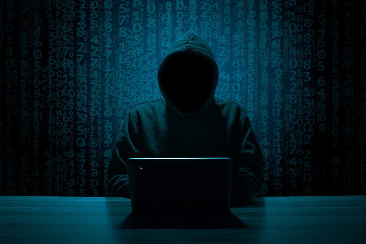 dark photo of a person wearing a black hoodie, with their face obscured, sitting at a laptop