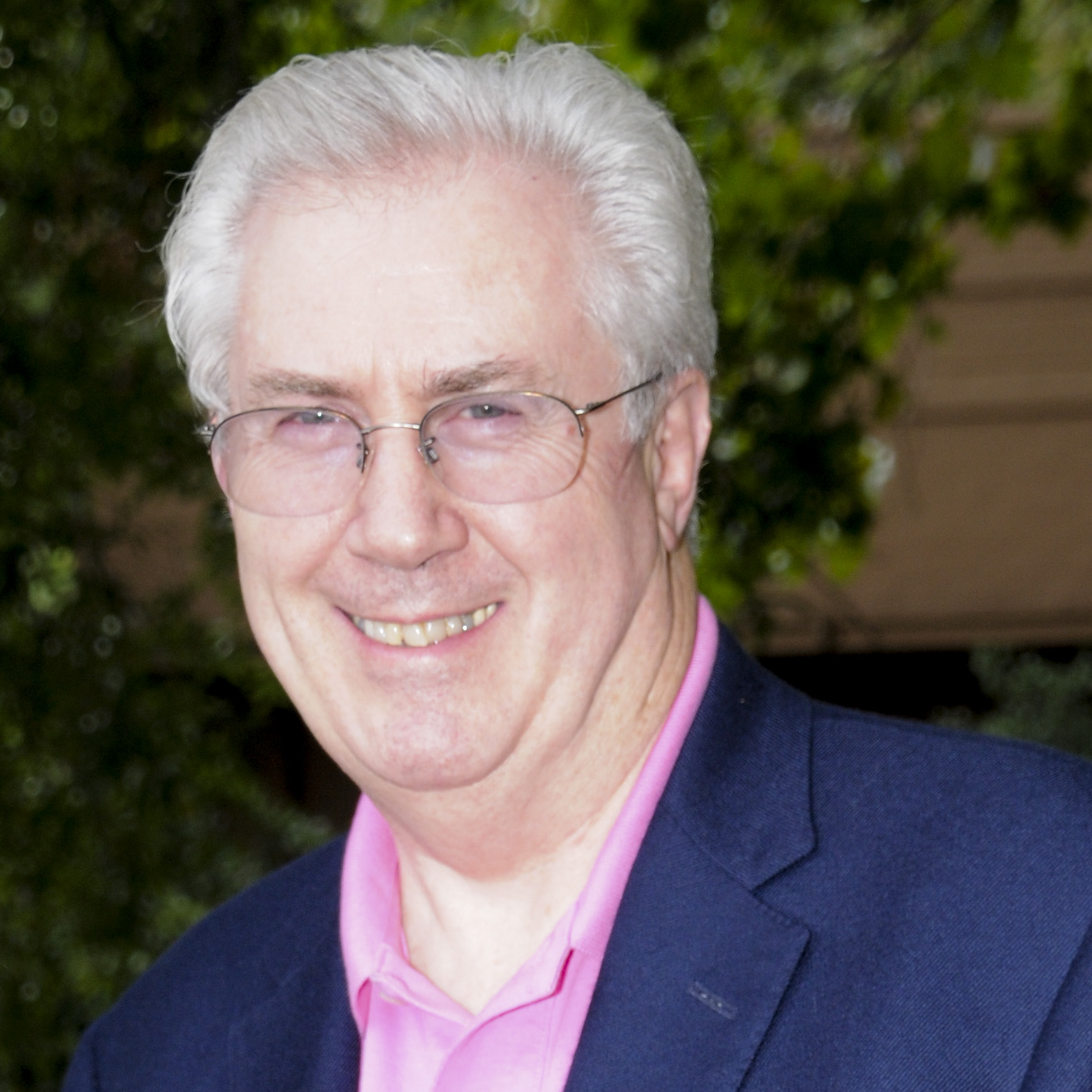 Headshot of a white man, with gray hair and glasses, smiling, wearing a pink shirt and blue sportcoat