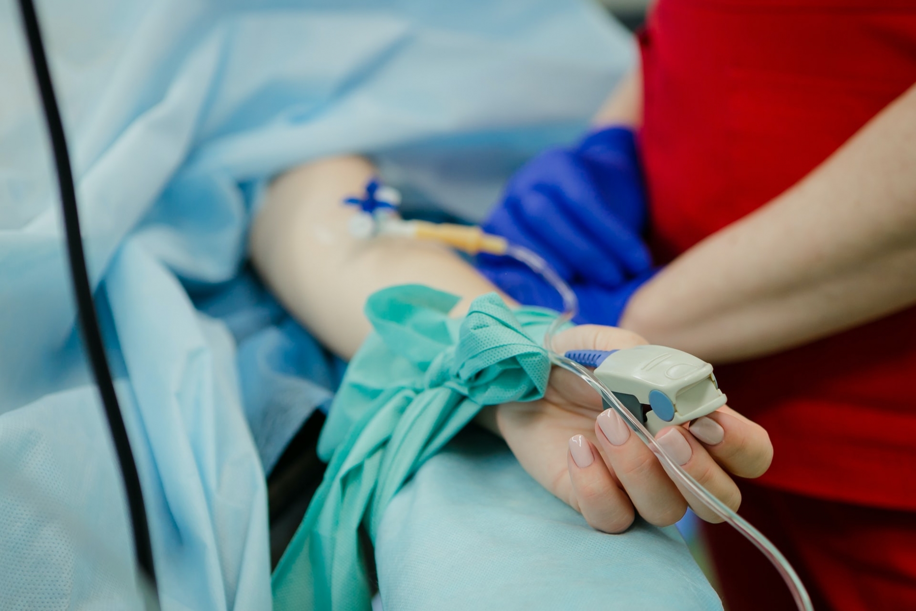 A woman's arm, extended from a hospital bed, with an inserted IV and a pulse sensor, while a person in red scrubs and blue latex gloves stands by her side.