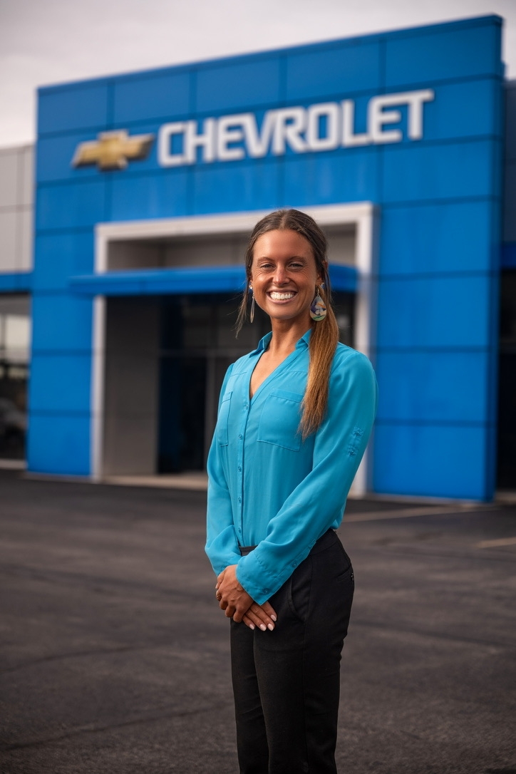 A blond woman, wearing a blue shirt and black pants, stands smiling in front of a Chevrolet dealership