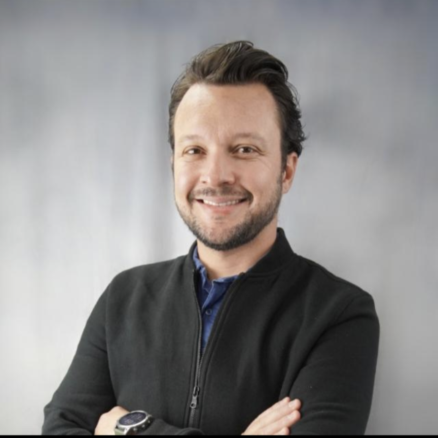 Headshot of a man with short brown hair, a short beard, wearing a black jacket, with his arms crossed and a smile on his face