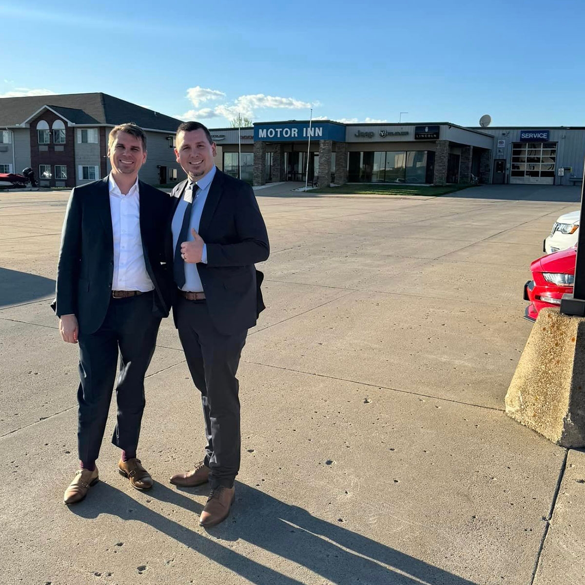 two men, wearing suits, standing in front of a car dealership
