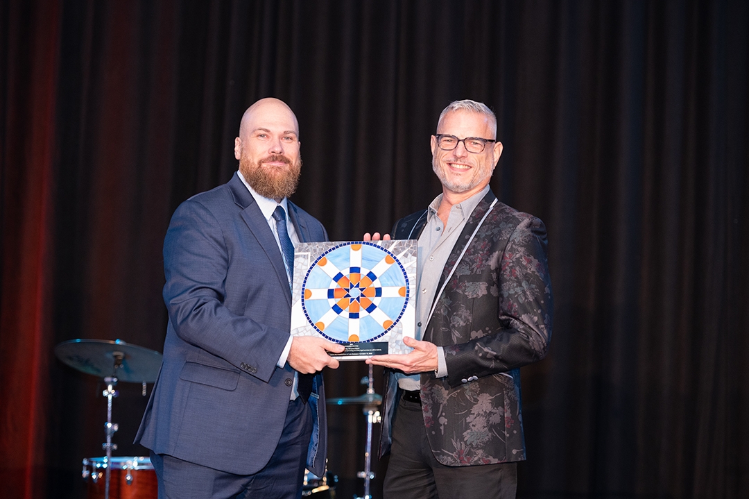 Photo of two men in suits, standing on stage, holding an award plaque