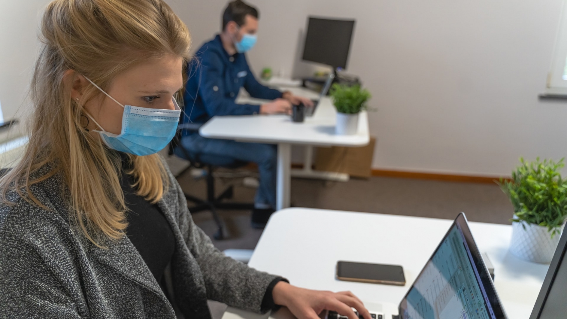 A woman and a man, wearing masks, working on computers at separate desks in an office