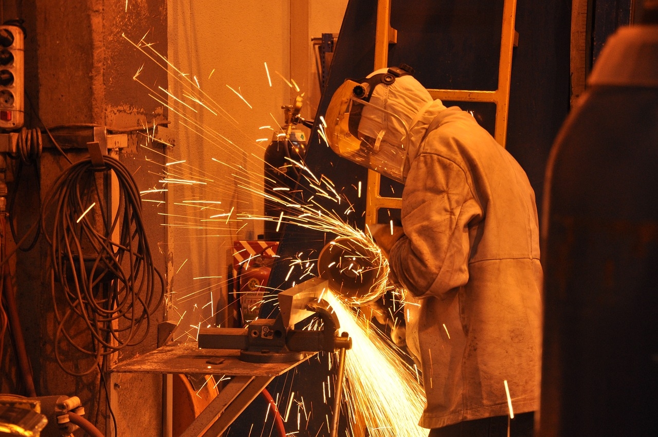 Person wearing a face shield and protective jacket, using a metal grinder, with sparks flying everywhere