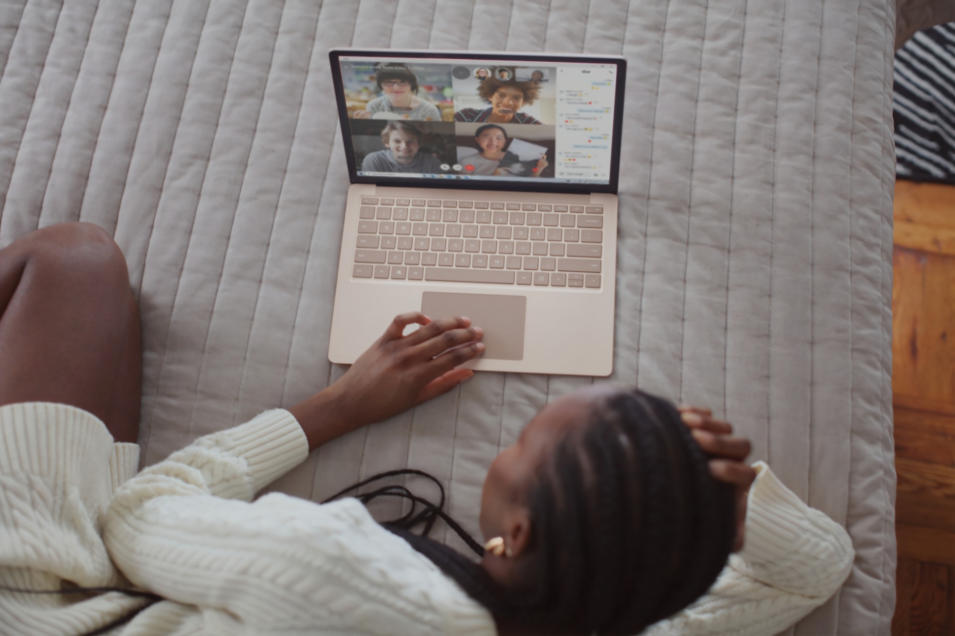 A woman lounges on a bed while video conferencing with four people on a notebook computer