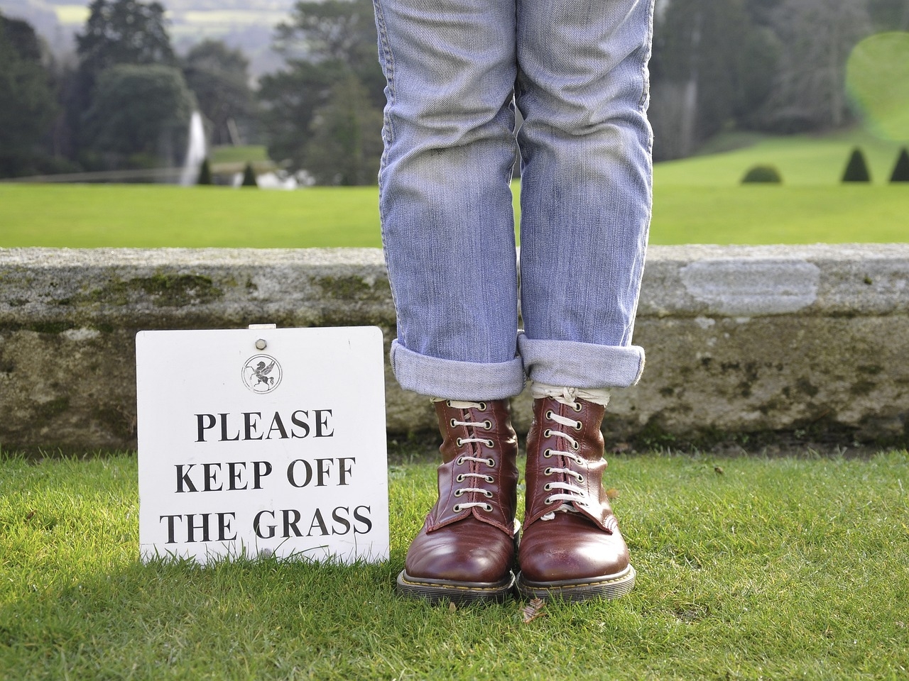 Photo of the boots and jean-covered legs of someone standing on a grass lawn, next to a sigbn that reads Please Keep Off the Grass