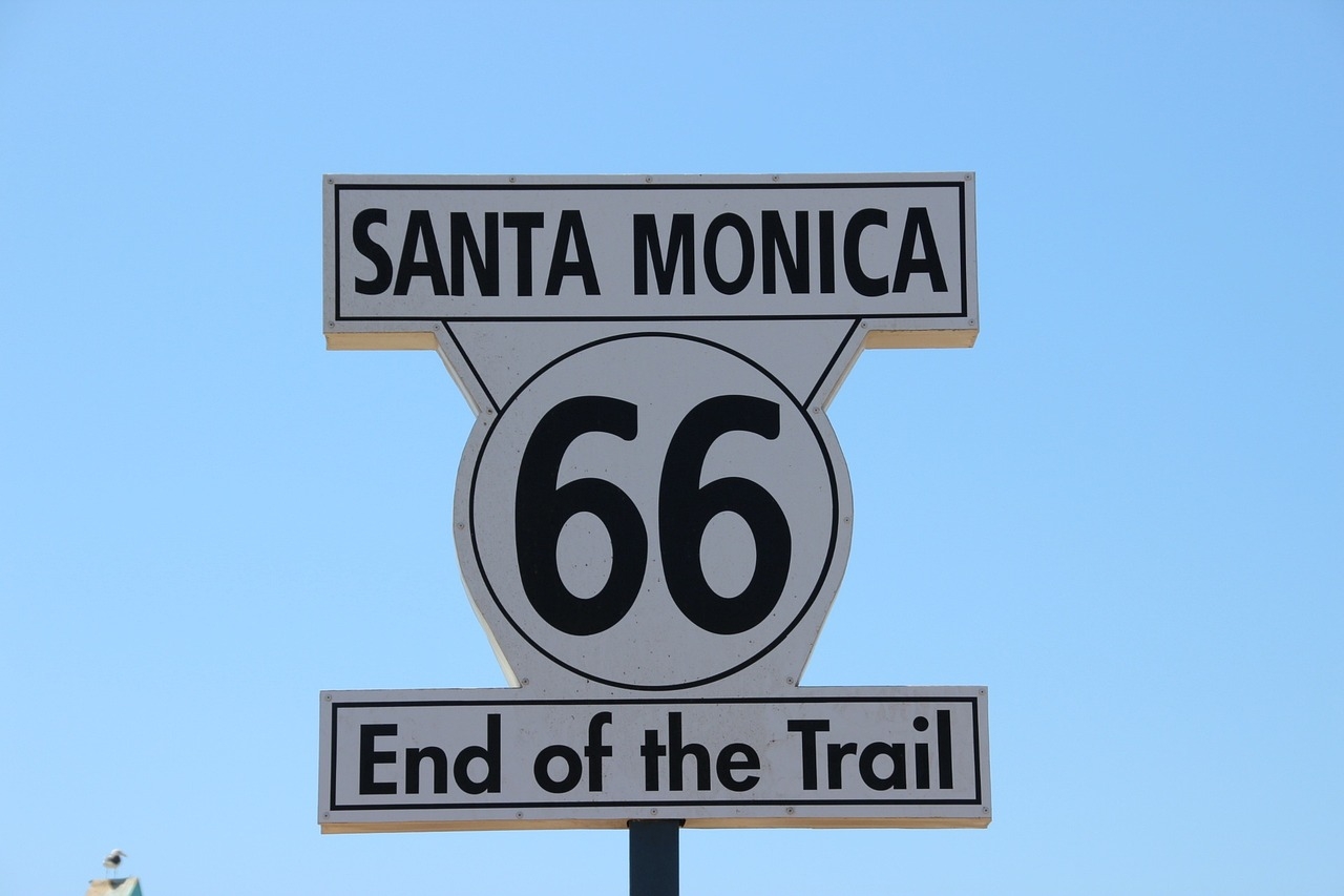 A white sign against a blue sky, with black text that reads Santa Monica 66 end of the trail