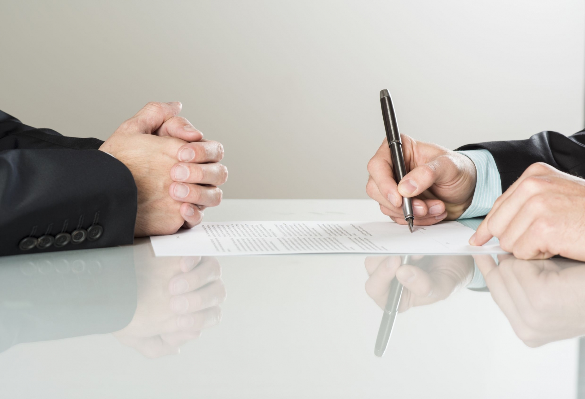 The hands of two men sitting across the table from one another, with one signing a piece of paper.