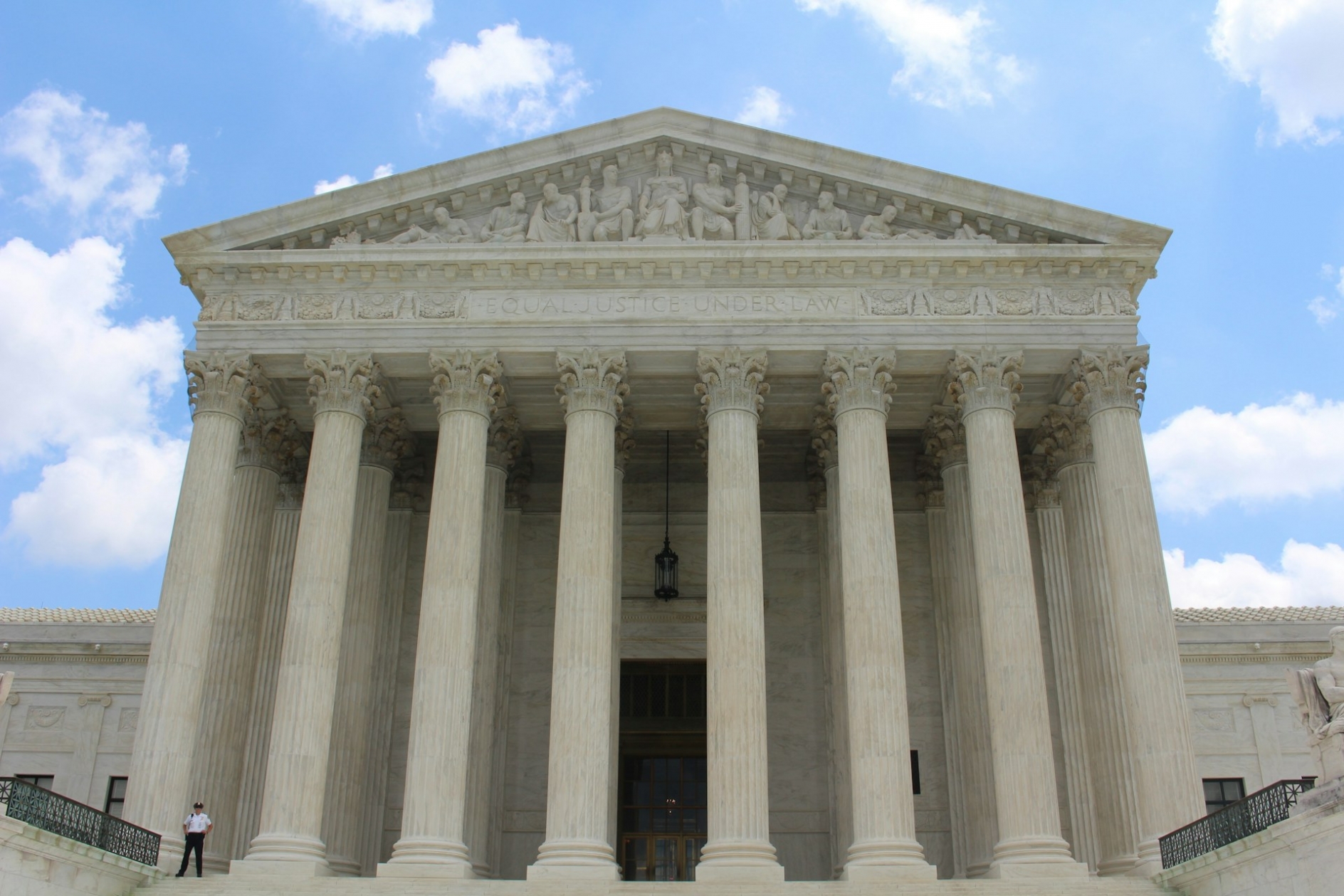 The front facade of the Supreme Court of the United States, under blue sky, with an officer standing guard