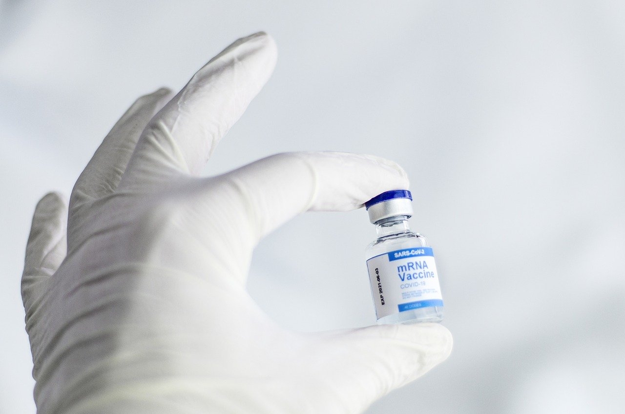 A left hand, wearing a white latex glove, and holding a vial of mRNA vaccine