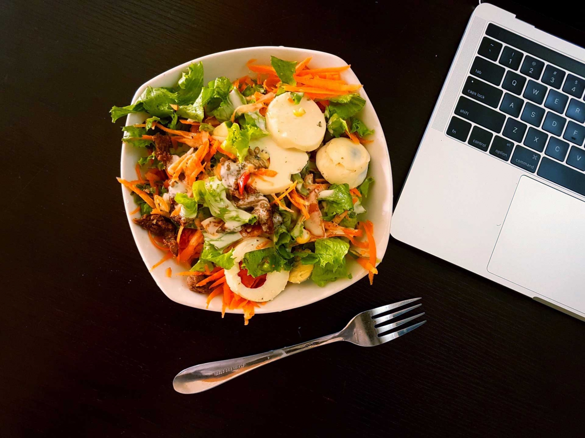 Salad in a bowl with a fork and an open laptop computer sitting next to it on the desk