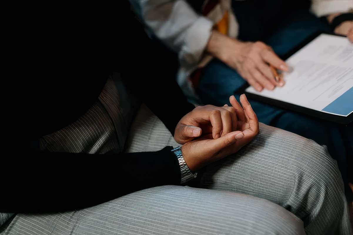 The hands of a black woman, lightly clasped in her lap, and the hands of a white woman, holding a clipboard and pen, reviewing paperwork together